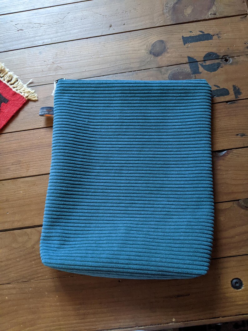 Peut inclure: Une pochette zipp&eacute;e en velours c&ocirc;tel&eacute; bleu sarcelle. La pochette est pos&eacute;e &agrave; plat sur une surface en bois.