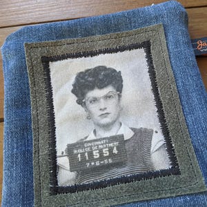 May include: A blue denim pouch with a green suede patch featuring a black and white photo of a woman with a mugshot style background. The photo includes the text "Cincinnati Police Department 11554 7-8-55".