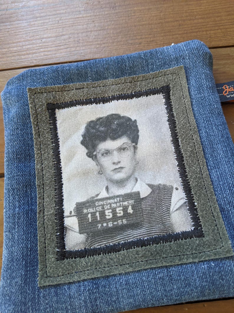 May include: A blue denim pouch with a green suede patch featuring a black and white photo of a woman with a mugshot style background. The photo includes the text "Cincinnati Police Department 11554 7-8-55".