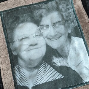 May include: Black and white photo of two women wearing glasses, smiling, and hugging. The photo is framed in a brown suede border with a green inner border.