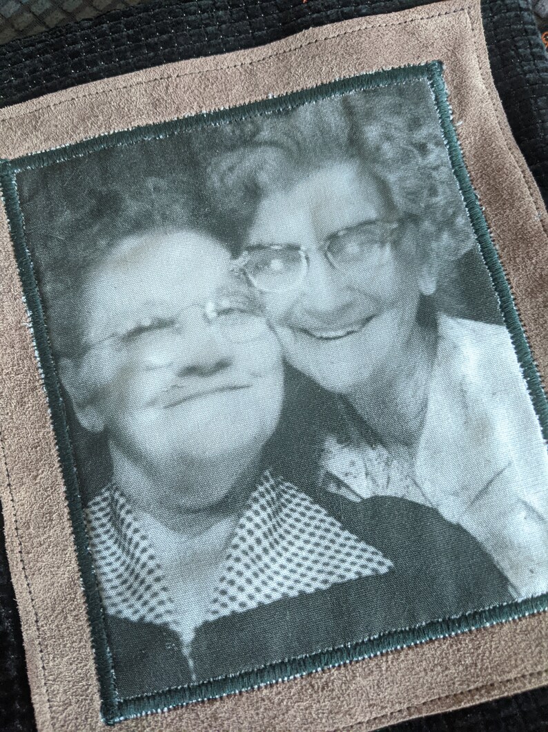 May include: Black and white photo of two women wearing glasses, smiling, and hugging. The photo is framed in a brown suede border with a green inner border.