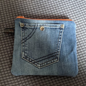 May include: A small blue denim pouch with a zipper closure and a large front pocket. The pouch has a light orange zipper pull and a silver button.