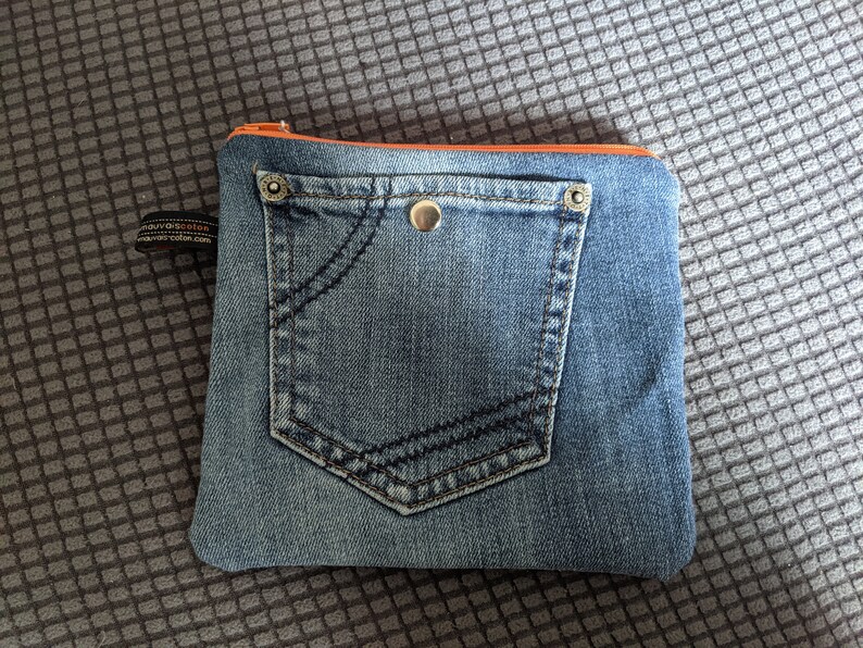 May include: A small blue denim pouch with a zipper closure and a large front pocket. The pouch has a light orange zipper pull and a silver button.