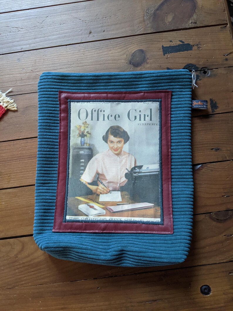 Peut inclure: Une pochette en velours c&ocirc;tel&eacute; bleu sarcelle avec une bordure rouge et blanche. La pochette pr&eacute;sente une couverture de magazine vintage avec le texte "Office Girl" et une photo d'une femme tapant sur un bureau.