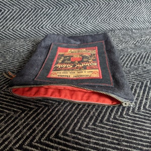 Puede incluir: Una bolsa de cremallera de denim con forro rojo. La bolsa presenta un parche cosido de estilo vintage con el texto "PARAMOUNT THEATRE" y "SHOCK SHOW" en letras rojas y negras.