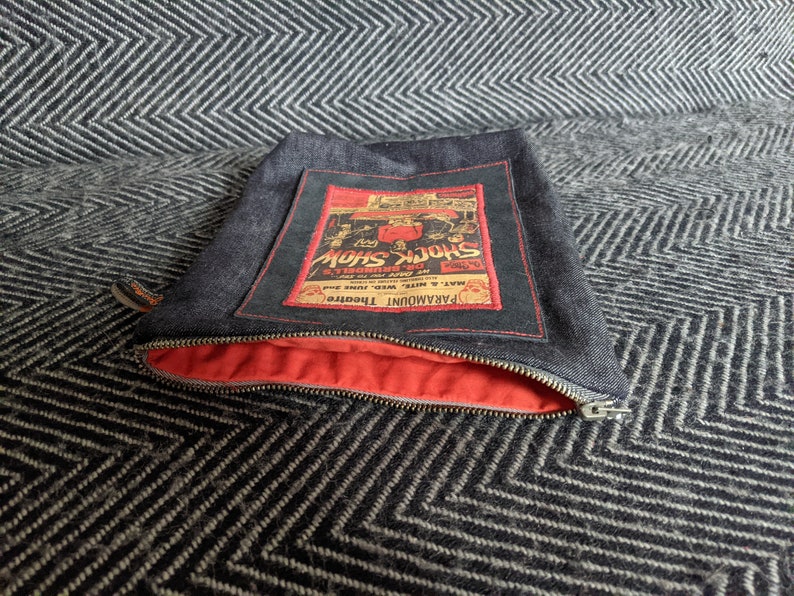 Puede incluir: Una bolsa de cremallera de denim con forro rojo. La bolsa presenta un parche cosido de estilo vintage con el texto "PARAMOUNT THEATRE" y "SHOCK SHOW" en letras rojas y negras.