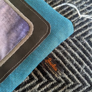 K&ouml;nnte beinhalten: Eine blaue und schwarze Stofftasche mit einem Lederklappenverschluss und einem grauen Stofffutter. Die Tasche hat ein schwarzes Etikett mit dem Text "Jackie filer" und "www.jackie-filer" in wei&szlig;.