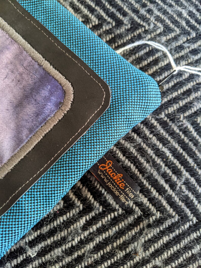 K&ouml;nnte beinhalten: Eine blaue und schwarze Stofftasche mit einem Lederklappenverschluss und einem grauen Stofffutter. Die Tasche hat ein schwarzes Etikett mit dem Text "Jackie filer" und "www.jackie-filer" in wei&szlig;.