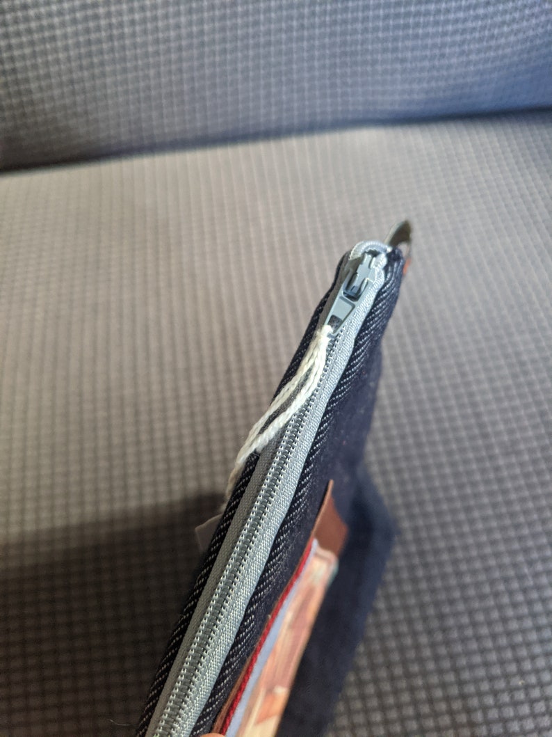 K&ouml;nnte beinhalten: Eine dunkelblaue, mit einem Rei&szlig;verschluss versehene Tasche aus Denim mit einem braunen Lederakzent und einem silbernen Rei&szlig;verschluss.