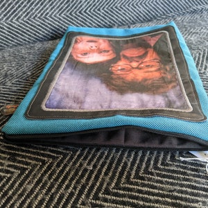 K&ouml;nnte beinhalten: Eine blaue Rei&szlig;verschlusstasche mit einem schwarzen Rei&szlig;verschluss und einem schwarzen und grauen Rand um ein Foto von zwei l&auml;chelnden Personen. Das Foto zeigt eine junge und eine &auml;ltere Person.