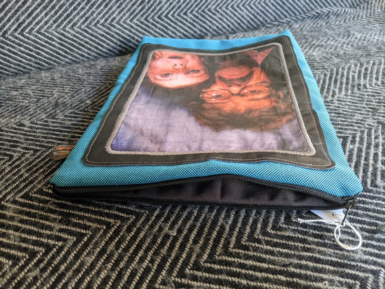 K&ouml;nnte beinhalten: Eine blaue Rei&szlig;verschlusstasche mit einem schwarzen Rei&szlig;verschluss und einem schwarzen und grauen Rand um ein Foto von zwei l&auml;chelnden Personen. Das Foto zeigt eine junge und eine &auml;ltere Person.