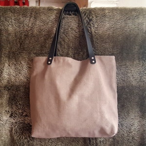 May include: A beige canvas tote bag with black leather handles and silver rivets. The bag has a simple design and is perfect for everyday use.