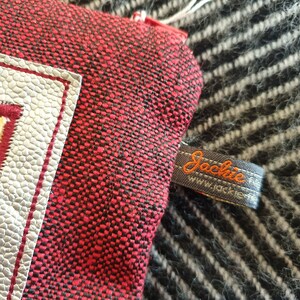 Op de afbeelding: Een rode en zwarte stoffen tas met een witte rand en een zilveren label. De tas heeft een zwart label met de tekst "Jackie www.jackie.de" in oranje draad.