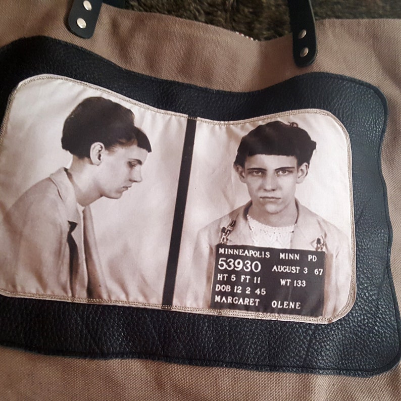 May include: A brown canvas tote bag with a black leather trim and a black and white photo of a woman's mugshot. The photo includes the text "MINNEAPOLIS MINN PD 53930 AUGUST 3 67 HT 5 FT 11 WT 133 DOB 12 2 45 MARGARET OLENE".