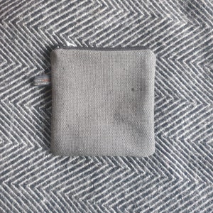 May include: A small, gray zippered pouch with a black and white textured fabric. The pouch is lying on a black and white patterned surface.