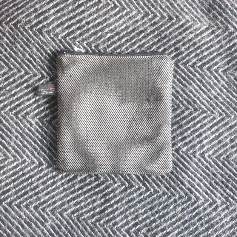 May include: A small, gray zippered pouch with a black and white textured fabric. The pouch is lying on a black and white patterned surface.