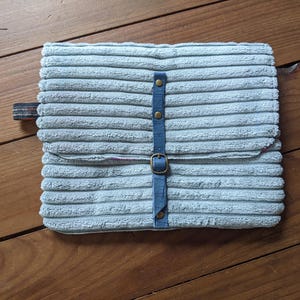 K&ouml;nnte beinhalten: Eine hellblaue Cord-Laptoptasche mit einem blauen Riemen und Messing-Schnallen.