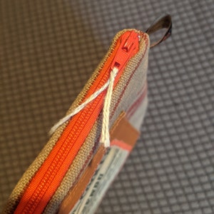 May include: A small zippered pouch with a red zipper and a brown and white striped fabric. The pouch has a brown leather patch with text on it.