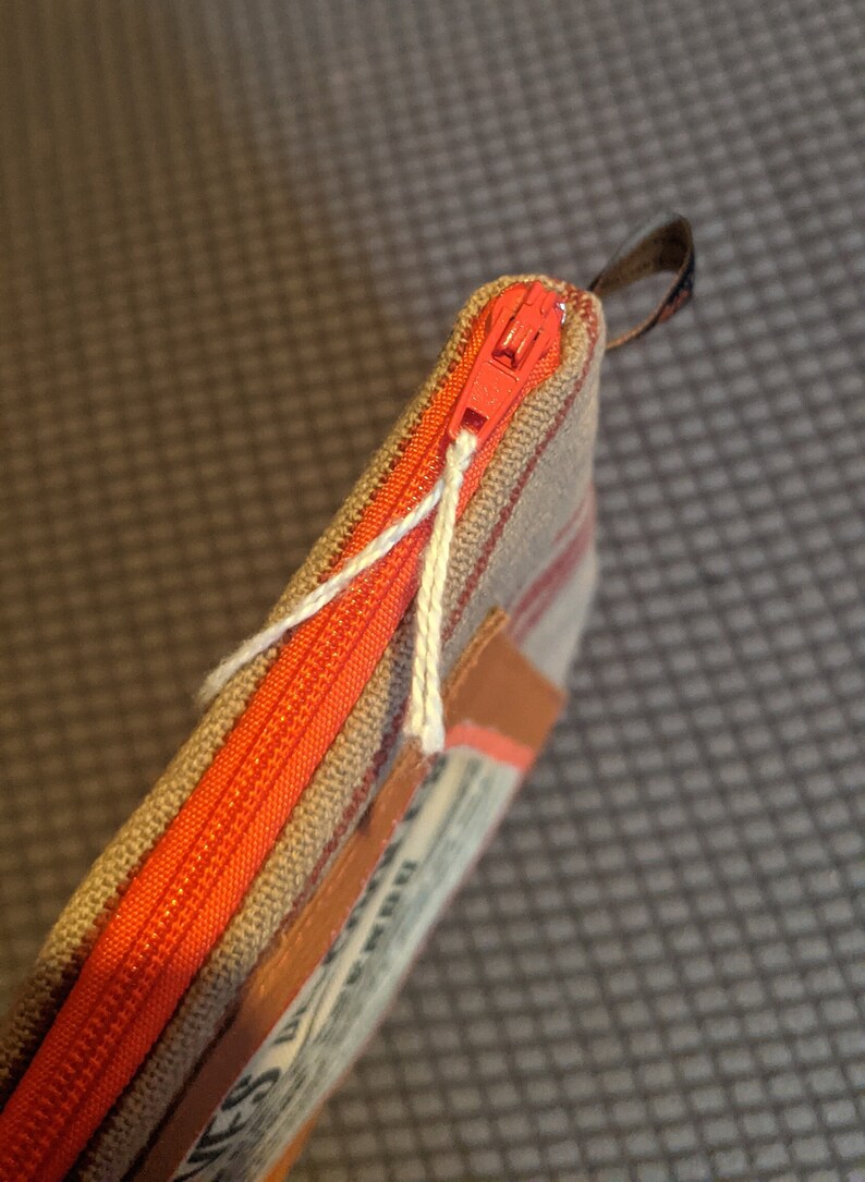 May include: A small zippered pouch with a red zipper and a brown and white striped fabric. The pouch has a brown leather patch with text on it.