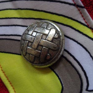 May include: A silver metal button with a woven design, set against a background of colourful striped fabric.