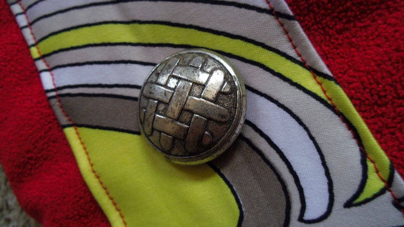 May include: A silver metal button with a woven design, set against a background of colourful striped fabric.