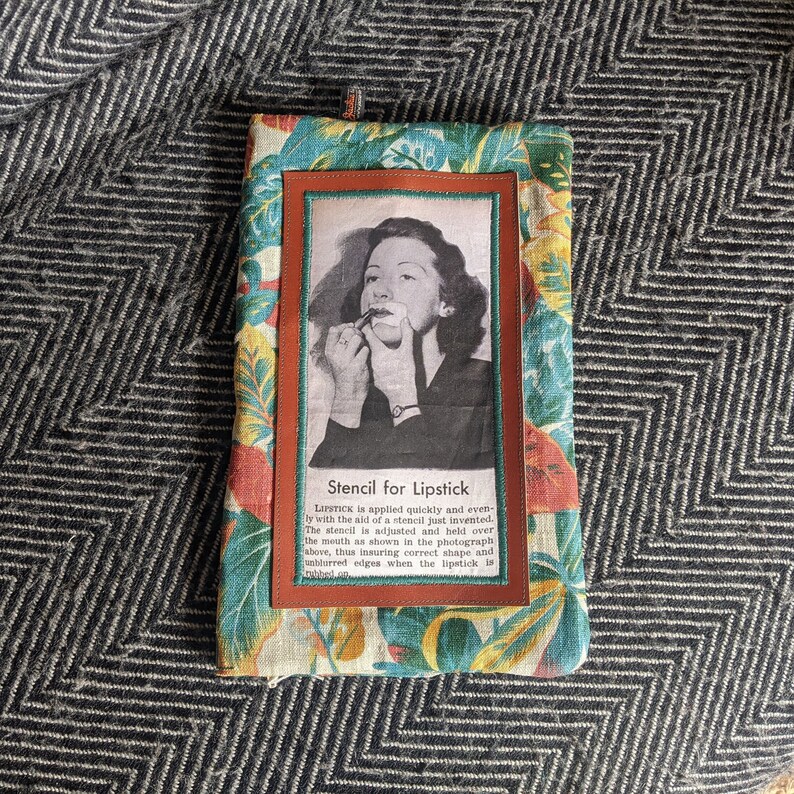 Pu&ograve; includere: Una pochette in tessuto vintage con stampa floreale. La pochette ha una patta in pelle marrone con un'immagine ritagliata di una donna che applica il rossetto. Il testo "Stencil for Lipstick" &egrave; stampato sulla patta. Il rossetto viene applicato rapidamente e uniformemente con l'aiuto di uno stencil appena inventato. Lo stencil viene regolato e tenuto sopra la bocca come mostrato nella fotografia sopra, garantendo cos&igrave; una forma corretta e bordi nitidi quando il rossetto viene applicato.