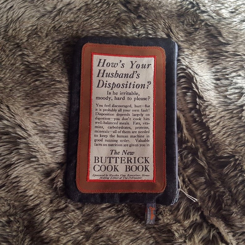 May include: A brown leather-bound cookbook with the title "The New Butterick Cook Book" and the text "How's Your Husband's Disposition? Is he irritable, moody, hard to please?"  The text explains that a husband's disposition depends on his diet and encourages readers to cook well-balanced meals.