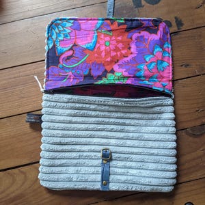 K&ouml;nnte beinhalten: Eine graue Cord-Tasche mit Rei&szlig;verschluss und einem floralen Stofffutter. Die Tasche hat einen blauen Lederriemen mit einer Metallschnalle.