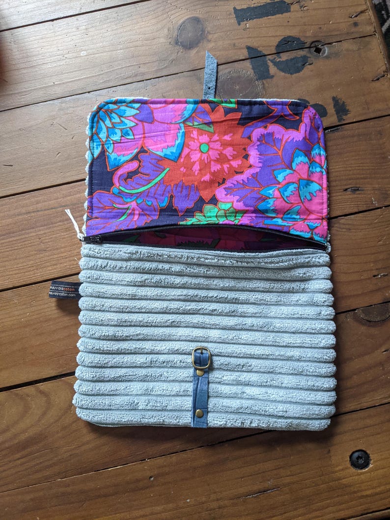 K&ouml;nnte beinhalten: Eine graue Cord-Tasche mit Rei&szlig;verschluss und einem floralen Stofffutter. Die Tasche hat einen blauen Lederriemen mit einer Metallschnalle.