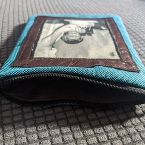 May include: A blue zippered pouch with a brown leather frame and a black and white photo of a person inside.