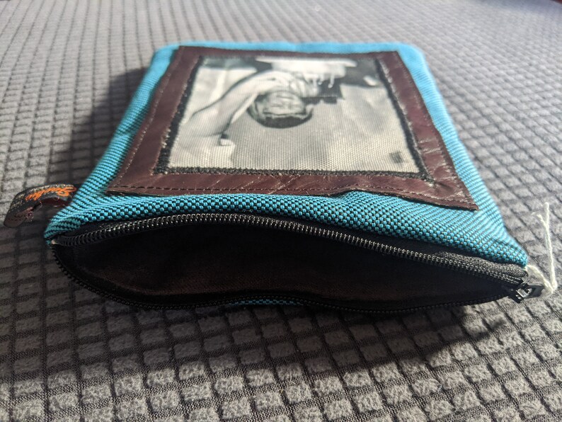 May include: A blue zippered pouch with a brown leather frame and a black and white photo of a person inside.