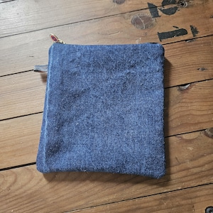 May include: A square, blue denim pouch with a gold zipper and a small leather pull tab. The pouch is made of a textured, faded denim material. The zipper pull has a decorative design.