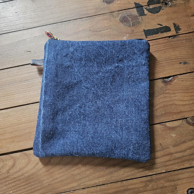 May include: A square, blue denim pouch with a gold zipper and a small leather pull tab. The pouch is made of a textured, faded denim material. The zipper pull has a decorative design.