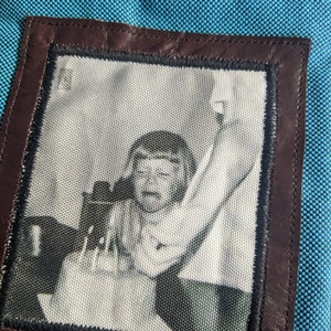 May include: A blue fabric pouch with a brown leather frame. The frame contains a black and white photo of a person with their arm raised.