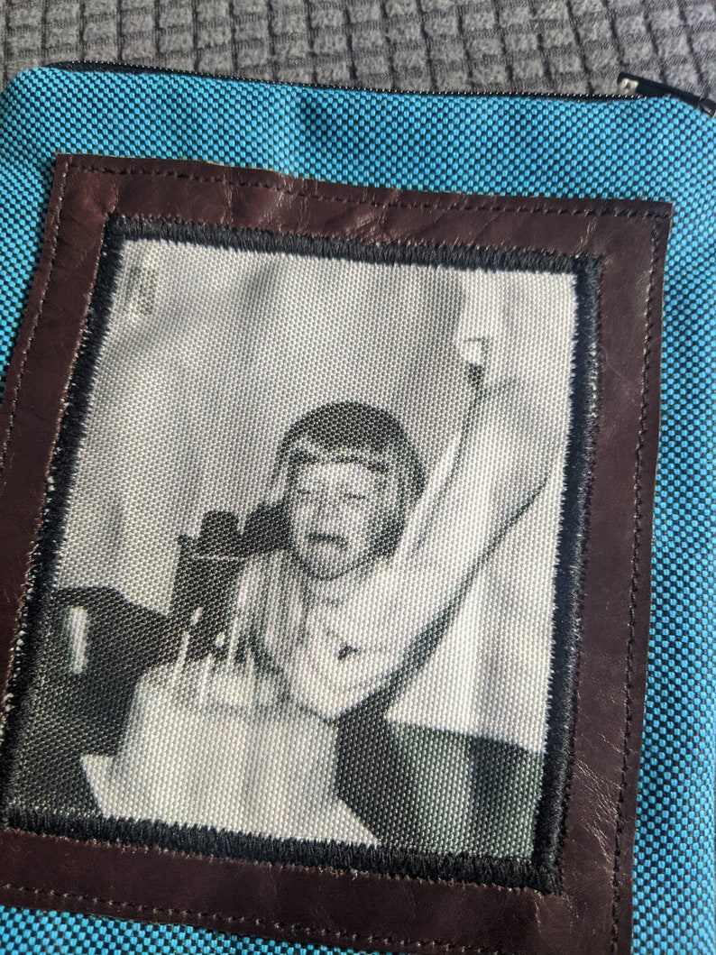 May include: A blue fabric pouch with a brown leather frame. The frame contains a black and white photo of a person with their arm raised.