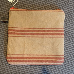May include: A beige zippered pouch with red stripes. The pouch is made of a woven fabric.
