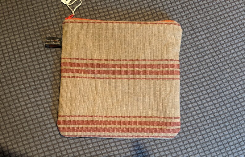 May include: A beige zippered pouch with red stripes. The pouch is made of a woven fabric.