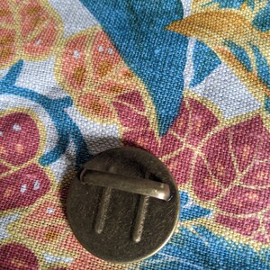 Pu&ograve; includere: Un bottone in metallo dorato con un design decorativo e un anello sul retro. Il bottone &egrave; su uno sfondo di tessuto floreale.