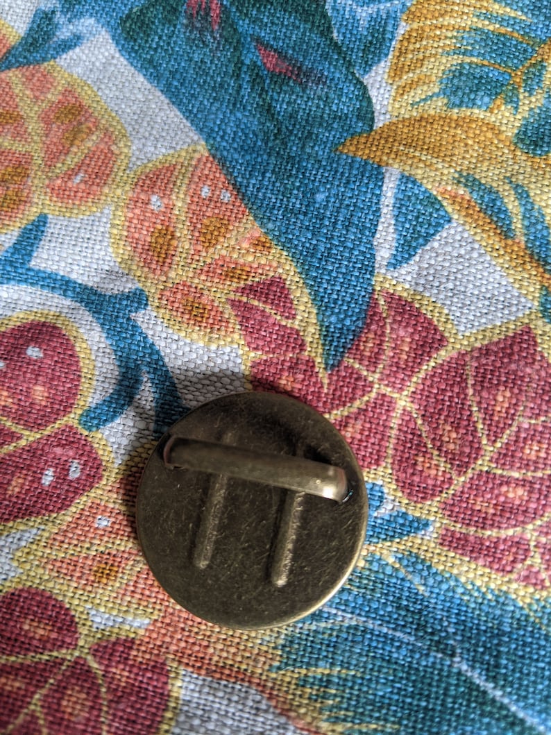 Pu&ograve; includere: Un bottone in metallo dorato con un design decorativo e un anello sul retro. Il bottone &egrave; su uno sfondo di tessuto floreale.