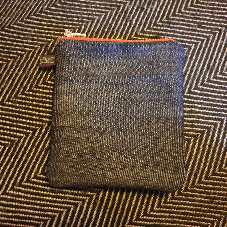 May include: A small, dark blue denim zippered pouch with an orange zipper pull. The pouch is lying on a black and white patterned surface.
