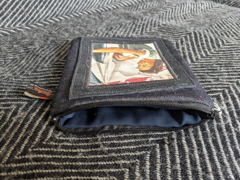 May include: A black denim zippered pouch with a rectangular window featuring a vintage photo of a woman with a red and white scarf. The pouch has a black zipper and a brown label with the word "Genuine" printed on it.