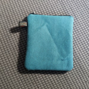 May include: A teal blue zippered pouch made of a textured fabric. The pouch is lying on a gray textured surface.