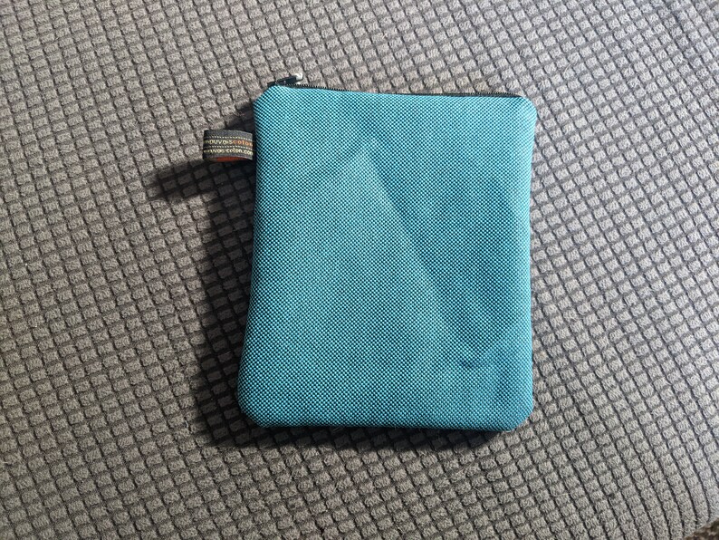 May include: A teal blue zippered pouch made of a textured fabric. The pouch is lying on a gray textured surface.