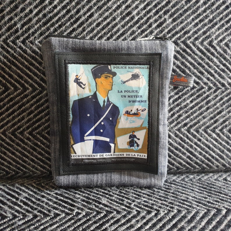May include: A gray zippered pouch with a black leather patch featuring a vintage French police recruitment poster. The poster depicts a police officer in uniform with the text "Police Nationale, La Police, Un Metier D'Homme, Recrutement de Gardiens de la Paix".