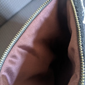 May include: A black zippered pouch with a brown fabric lining. The zipper is gold colored.