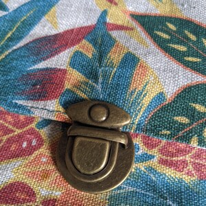 Pu&ograve; includere: Un fermo in ottone su una borsa di tessuto con stampa floreale tropicale. Il fermo &egrave; rotondo con un cerchio rialzato al centro e una barra curva.