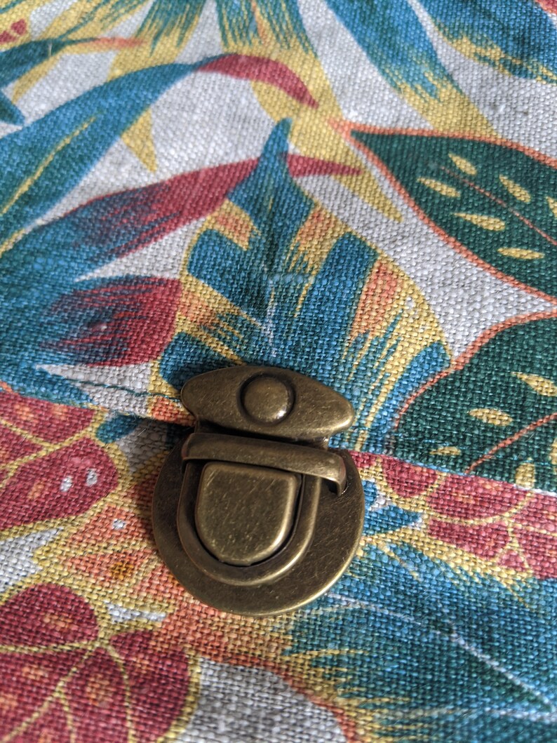 Pu&ograve; includere: Un fermo in ottone su una borsa di tessuto con stampa floreale tropicale. Il fermo &egrave; rotondo con un cerchio rialzato al centro e una barra curva.