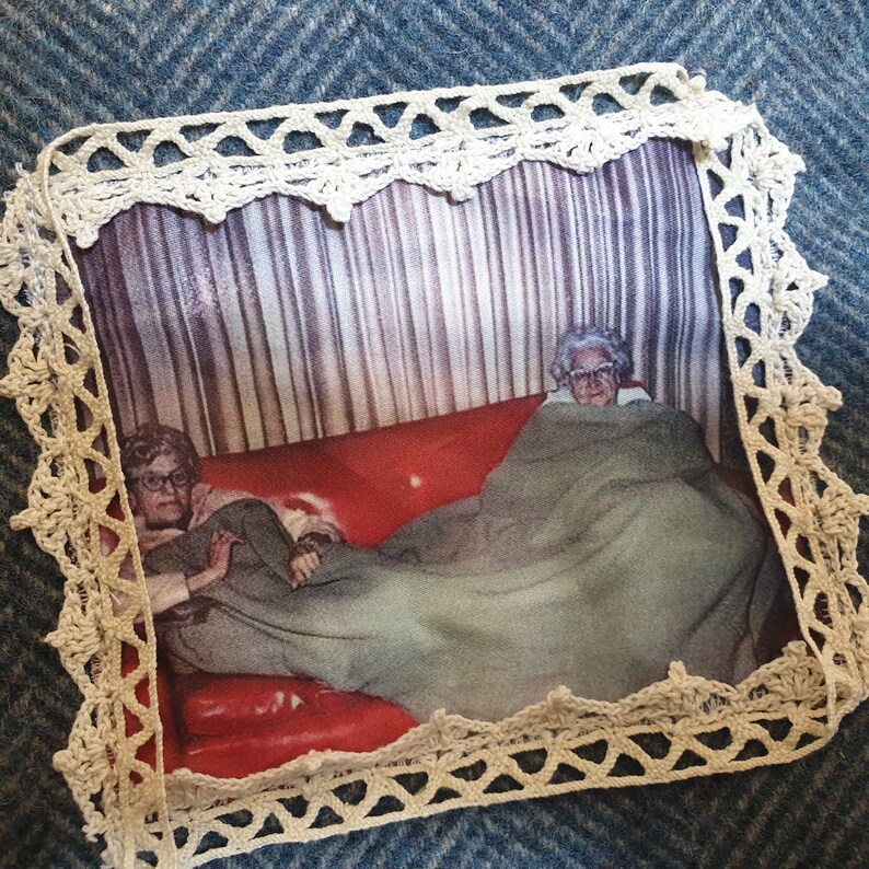 May include: A vintage photograph of two older women sitting on a red couch under a blanket. The photo is framed with white crocheted lace.