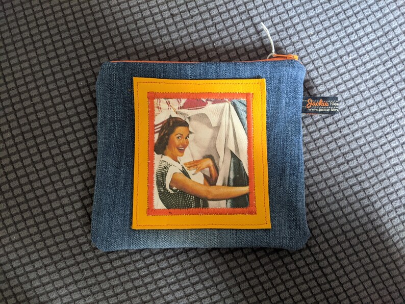 May include: A blue denim zippered pouch with a yellow and orange frame around a vintage image of a woman holding a sheet. The pouch has a small label that reads "Jackie 100% cotton Jackie's Library".