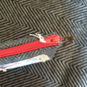 May include: Close-up of a red zipper on a gray and blue tweed fabric pouch. The zipper is partially open and the fabric is textured.